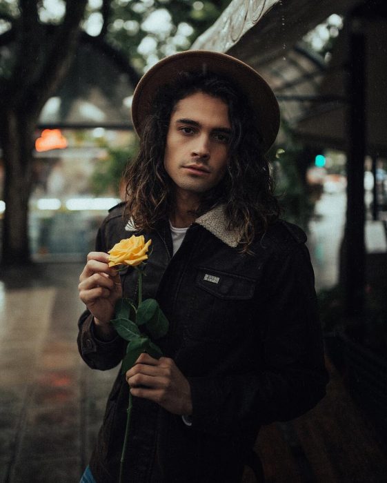 Catarino, hombre boho, con cabello largo y ondulado usando sombrero, con una rosa amarilla en las manos