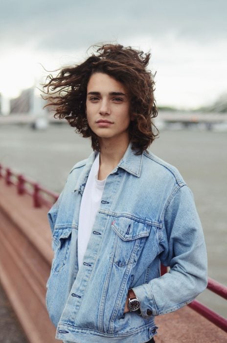 Matthew Clavane, modelo joven, chico con cabello largo a los hombros y ondulado, con chaqueta denim y manos en los bolsillos