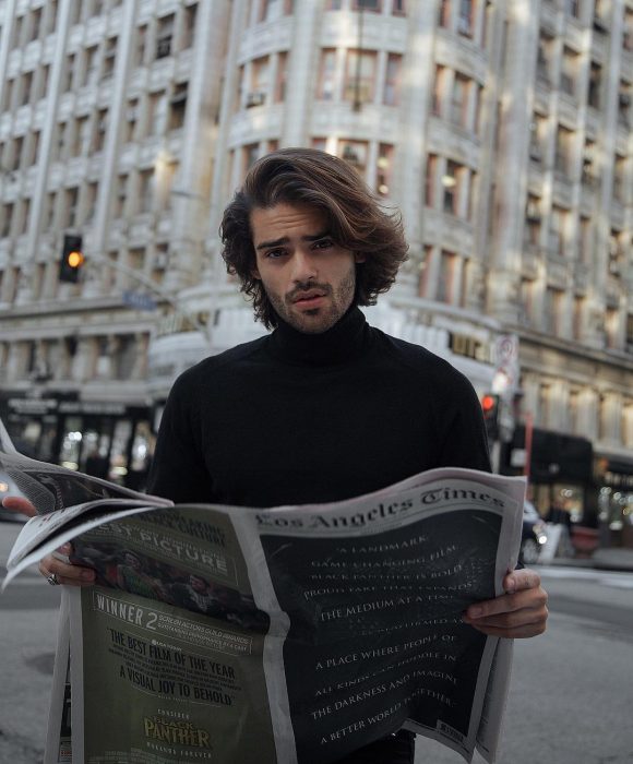 Renan Pacheco, modelo, hombre con el cabello castaño hasta los hombres, con bigote y barba corta, leyendo el periódico en la calle