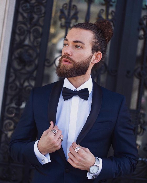 Hank, hombre de cabello castaño y largo recogdo en un man bun, con perforación en la nariz y traje de gala color azul marino y moño