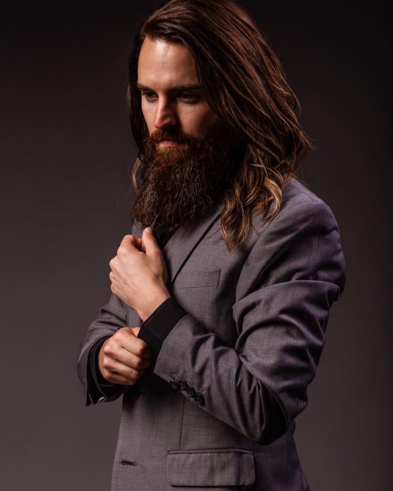 Simon, hombre de cabello largo y castaño, con bigote y barba larga, usando traje gris