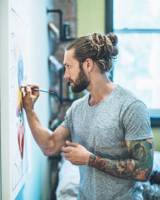 William Tyler, hombre de cabello rubio, largo y peinado man bun, con tatuajes en los brazos pintando un mural en la pared