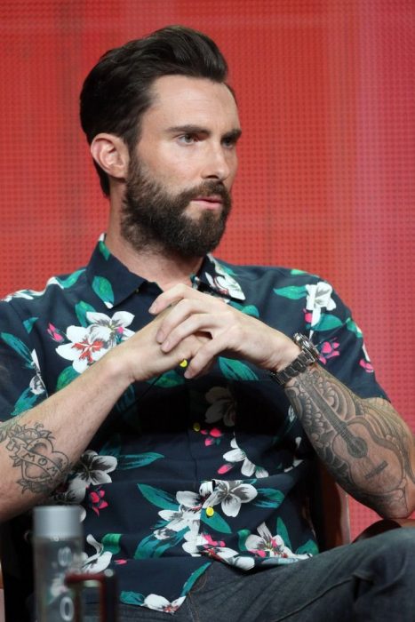 Vocalista de Maroon 5, Adam Levine, hombre sentado, con cabello corto y barba y bigotes, brazos tatuados usando una playera con flores blancas