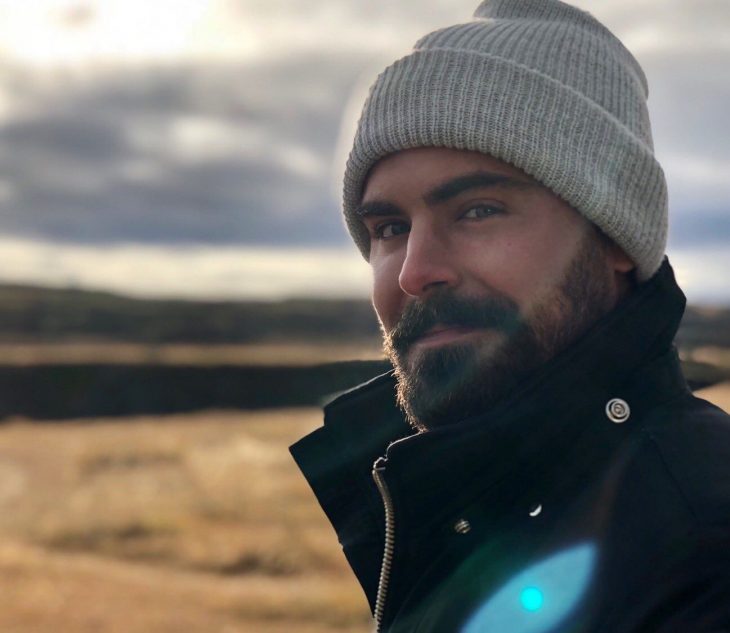 Actor Zac Efron, hombre con barba y bigote tupido, cejas pobladas, con gorro gris para el frío y chamarra