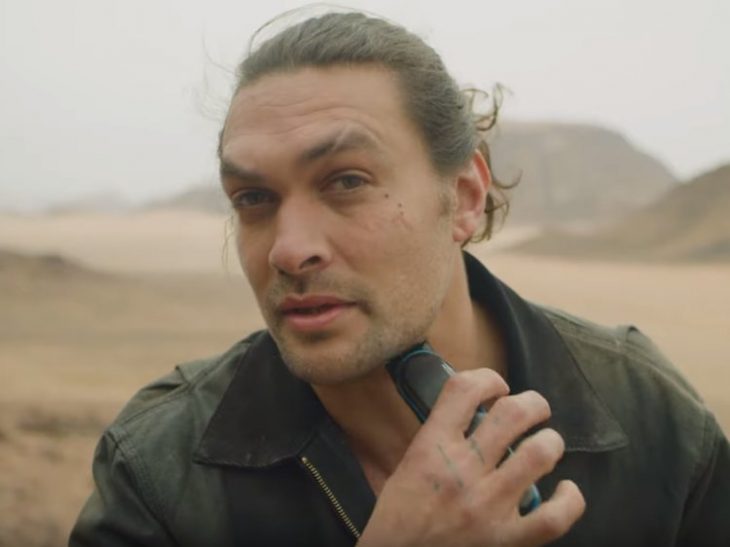 Jason Momoa a mitad de un bosque desertico, afeitando su barba, mientras ve a la cámara