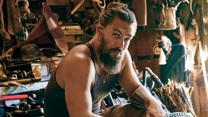 Jason Momoa con barba abultada, camisa sin mangas, sentado en una banca, dentro de una habitación de madera, antes de quitarse la barba