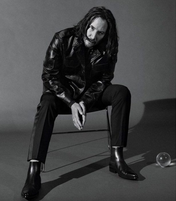 Keanu Revees sentado en una silla con las manos unidas, la cabeza inclinada, cubriendo su rostro con cabello largo, GQ, Jhon Wick: Parabellum