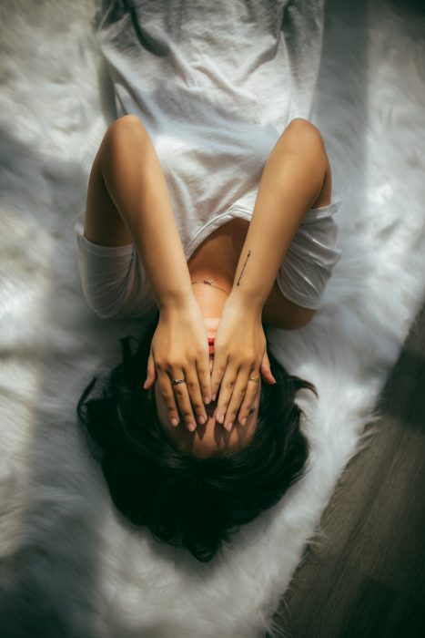 Chica recostada en una cama con sabanas blancas, cubriendo sus ojos con las palmas de sus manos, usando camisa blanca