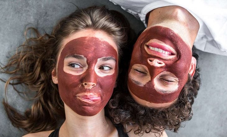 Pareja de novios recostados sobre el piso usando mascarilla de arcilla roja para las impurezas