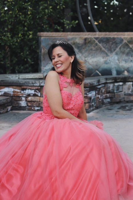 Mujer sentada de lado, mirando sobre su hombre, usando tiara en su cabeza, cabello rizado, sonriendo, llevando vestido ampon color coral, posando para una foto de XV años