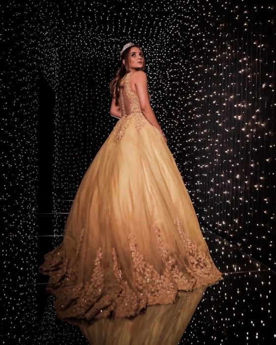 Chica usando vestido dorado posando en un cuarto cubierto con luces doradas, mirando de lado sobre su hombro, llevando tiara de piedras doradas, con peinado rizado