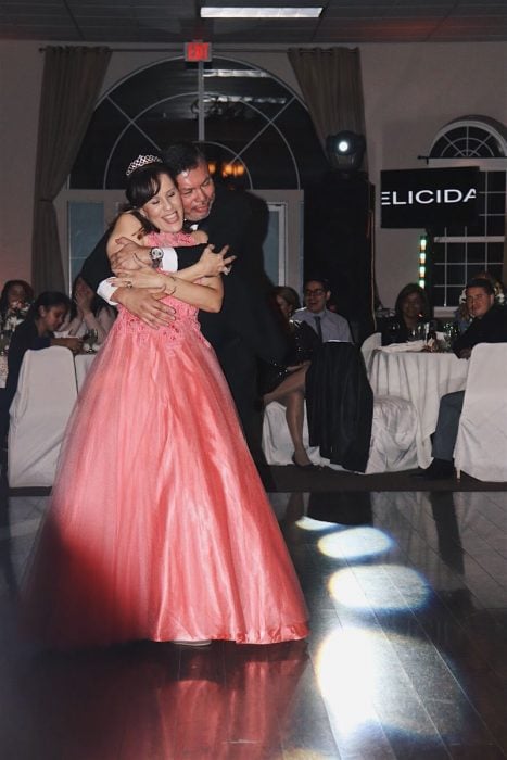 Hombre abrazando por la espalda a una mujer en una fiesta de XV años, después de bailar el vals, mujer llevando vestido color coral , ampon , tiara, hombre con traje sastre en tono negro y corbatín 
