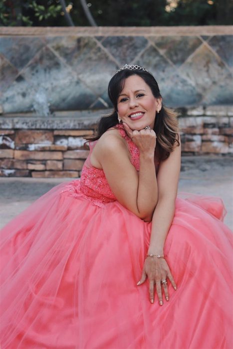 Mujer adulta usando vestido de quinceañera color coral, sentada en unas escaleras de asfalto, posando con su rostro recargado en su mano derecha