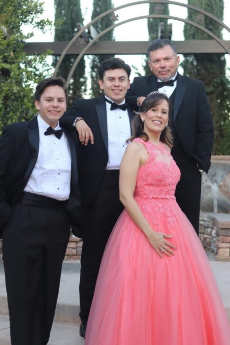 Familia posando frente a unas escaleras de asfalto para una foto del recuerdo, mujer usando vestido de quinceañera color coral, llevando tiara plateada, con el brazo en la cintura, hombres llevando traje sastre en colore negro con corbatín, con el brazo uno encima del hombro del otro