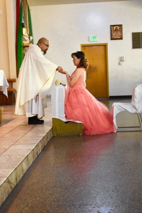 Mujer dentro de una iglesia, hincada frente a un sacerdote, tomando las manos de él, mirando al piso, hombre con sotan, agachado, entregando una ostia a una mujer durante una misa de celebración de xv años