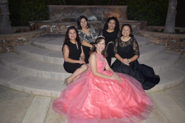 grupo de mujeres sentadas al filo de unas escaleras de aslfato, posando para una foto de recuerdo de una fiesta de xv años, son la spiernas cruzadas, llevando vestidos de color negro, con detalles en canaje, mujer al centro llevando vestido ampon de color coral estilo quinceañera