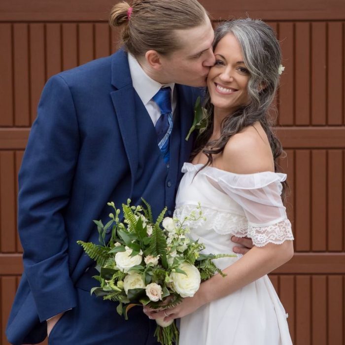 Pareja de esposos abrazados, mujer con cabello canoso, vestido blanco de novia y ramo de flores blancas, hombre de traje azul con cabello rubio, largo agarrado en una coleta