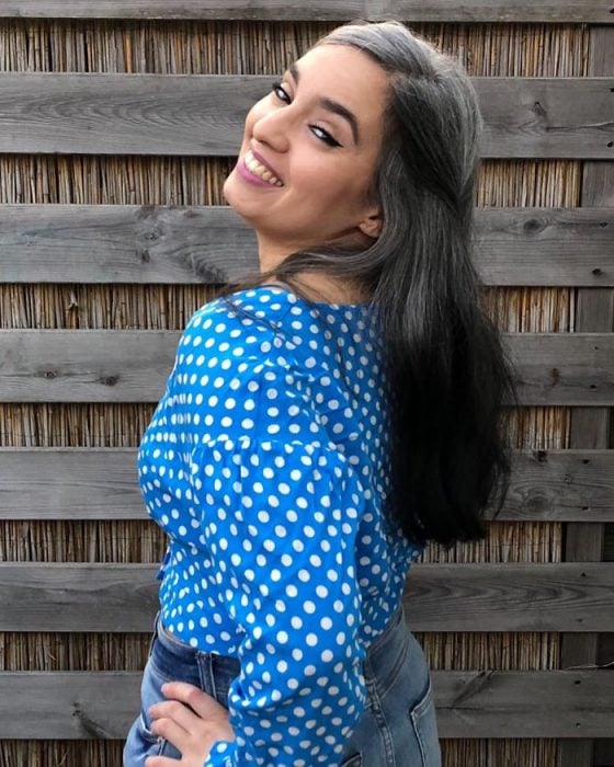 Chica de cabello largo y canoso posando para fotografía y sonriendo, blusa azul con puntos blancos