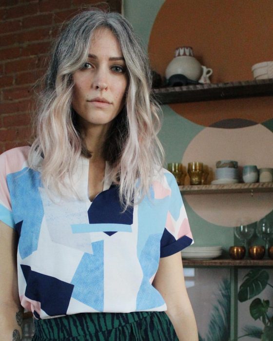 Mujer joven con cabello canoso ondulado, ojos grises, con blusa de patrones geométricos azul