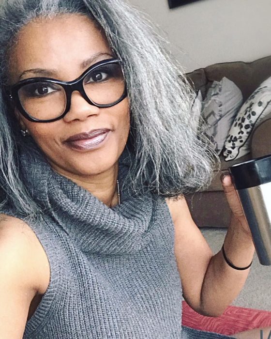 Mujer morena, afroamericana usando lentes y bebiendo café de un termo, blusa tejida gris y cabello corto con canas