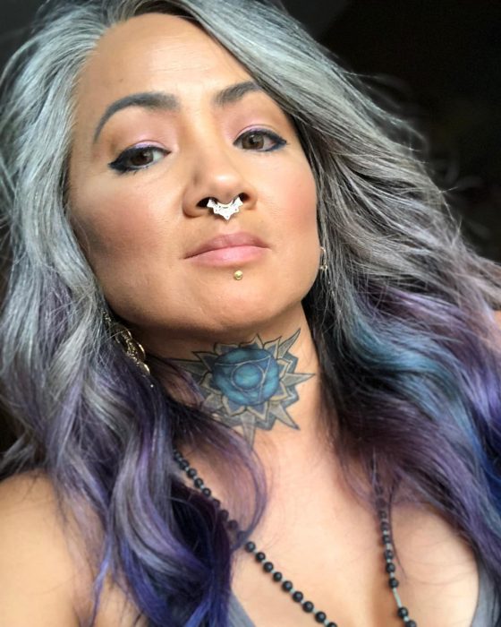 Mujer con tatuaje de flor de loto en el cuello, piercing en el septum y cabello colo gris y morado