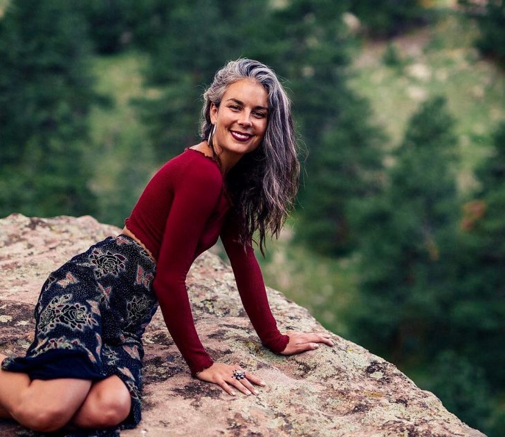 Mujer joven sonriendo, de cabello largo, ondulado y canoso, sentada en una roca con blusa roja y falda