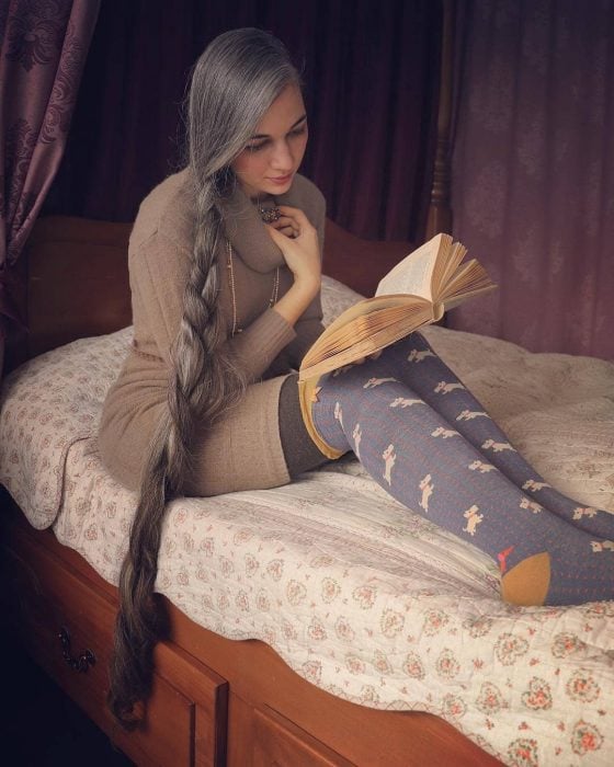Mujer joven con cabello canoso, muy largo y trenzado hacia un lado, leyendo un libro en la cama con calcetas largas azules con figuras de perros