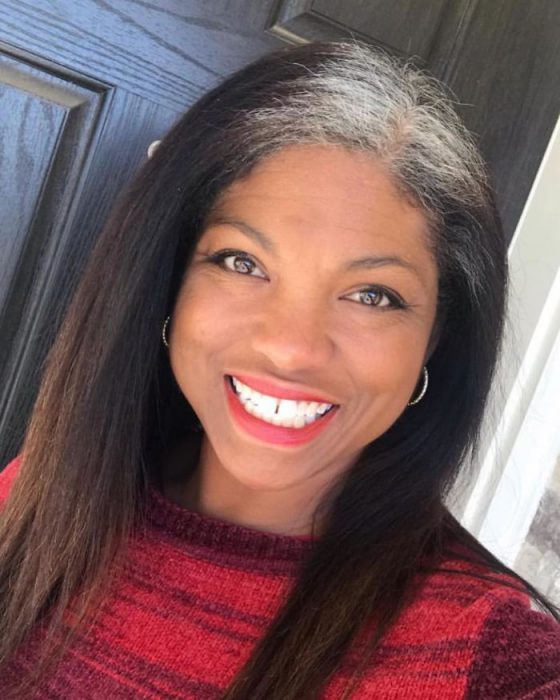 Mujer morena afroamericana, con cabello castaño y raíces blancas, sonriendo con los dientes separados