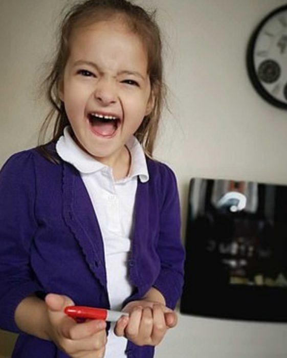 Lily Schooley, niña pequeña con uniforme escolar sosteniendo un plumón rojo y riéndose