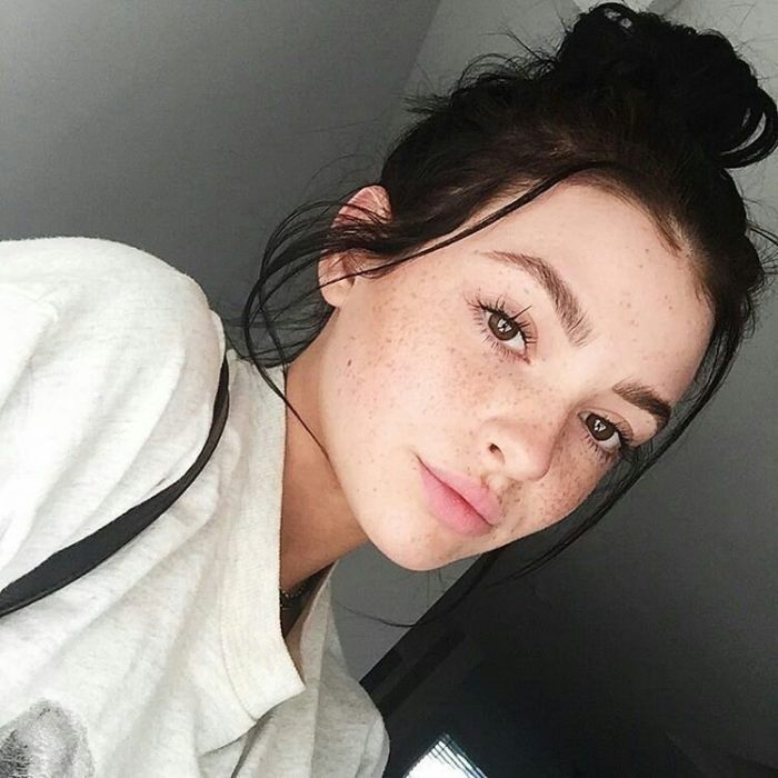 Chica con pecas en el rostros, peinado bun, sudadera blanca, sonriendo ligeramente, dentro de una habitación gris, tomando una selfie