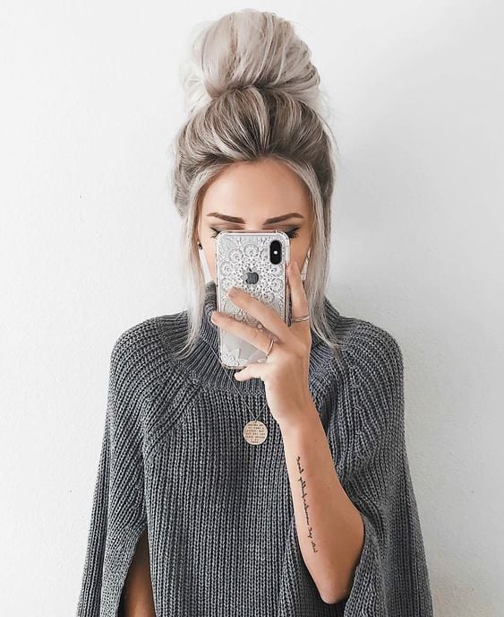 Chica tomándose una fotografía con su celular, con suéter gris y un peinado de moño alto despeinado