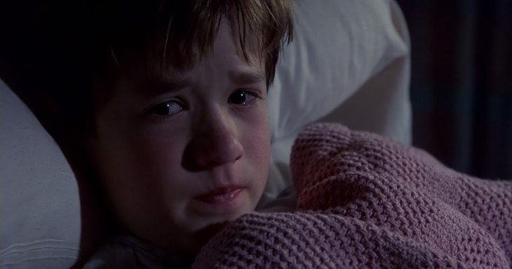 Niño recostado en su cama, cubriéndose con una manta de color rosa tejida, escena de la película El sexto sentido, Haley Joel Osment