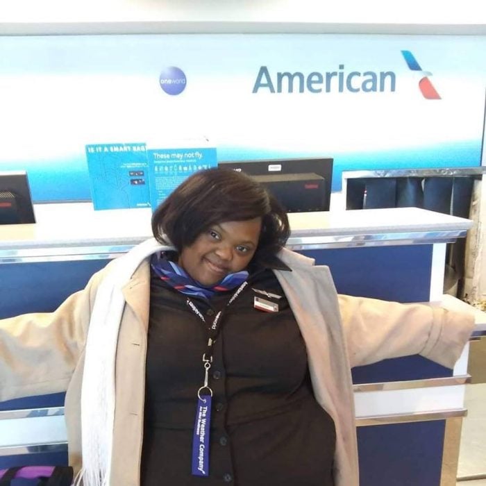 Shantell Pooser vestida de azafata, recargada en un mostrador de documentación del aeropuerto, mirando hacia la cámara y sonriendo