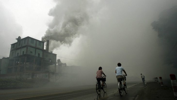 Boys pass by bicycle near a factory with polluting waste