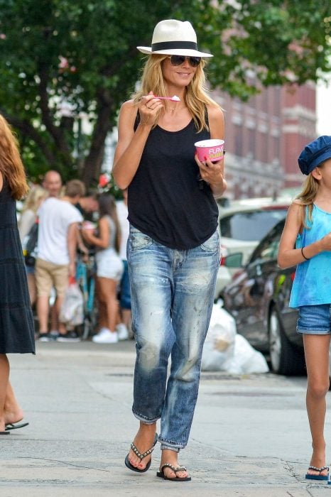 Girl walking down the street eating ice cream while wearing jeans, a black blouse, sandals and hat 