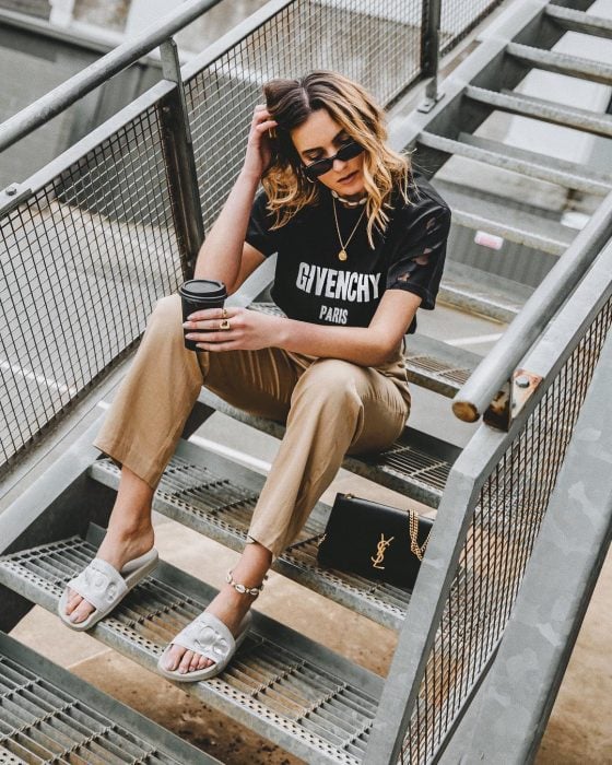 Girl wearing white sandals, brown pants and black blouse while sitting on some stairs drinking coffee 