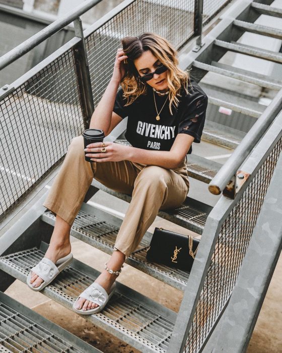 Chica usando unas sandalias de color blanco, pantalón café y blusa negra mientras está sentada en unas escaleras tomando café 