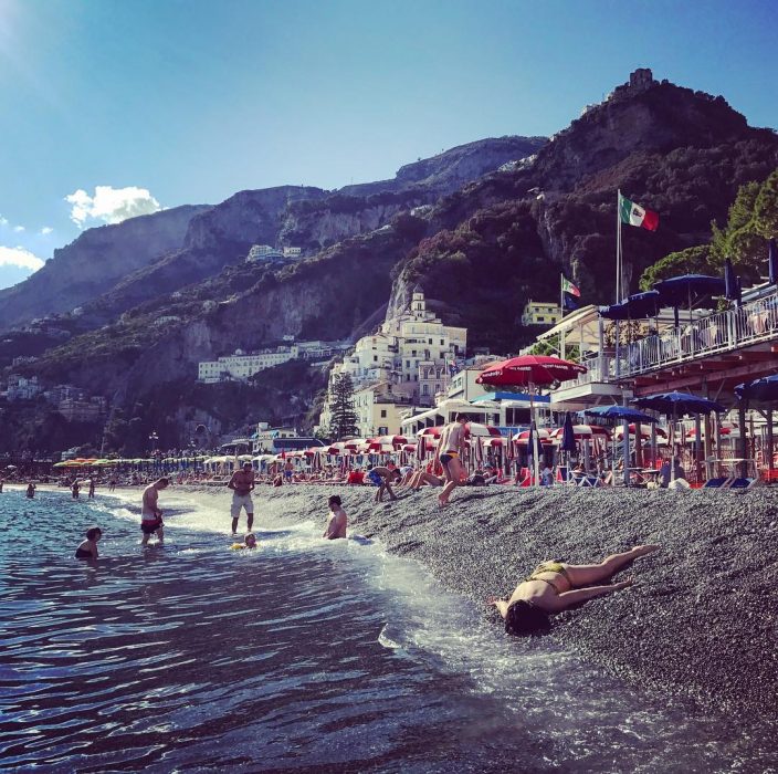 STEFDIES, la chica que se ha vuelto viral por sus antiselfies, selfie de mujer en la costa junto al mar en playa Amalfitana en Italia, con montañas en el fondo y turistas