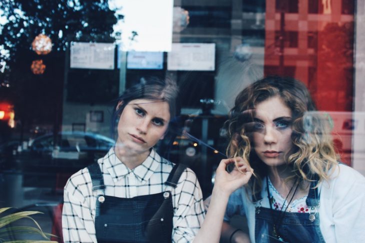 Tipo de amiga Libra, amigas llevando outfits iguales con overoles de mezclilla, mirando a atrvés de la ventana de una lavandería 