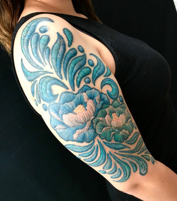 Chica parada de perfil mostrando su brazo derecho con un tatuaje de guías en color azul con efecto bordado 
