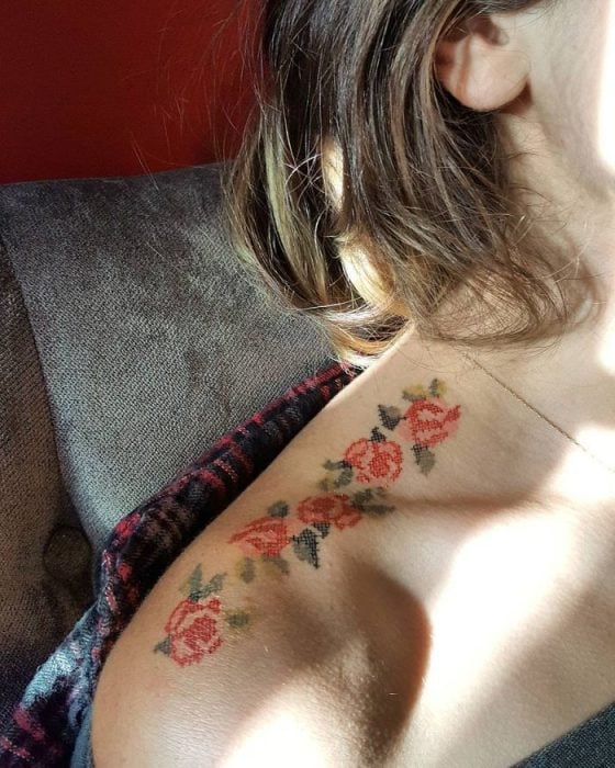 Chica recostada en un sofá, mostrando su hombro descubierto para enseñar su tatuaje de guía de rosas con efecto bordado en punto de cruz