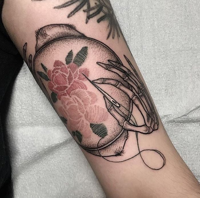 Persona mostrando su antebrazo tatuado con el diseño de una mano bordando una rosa en un telar con efecto bordado 