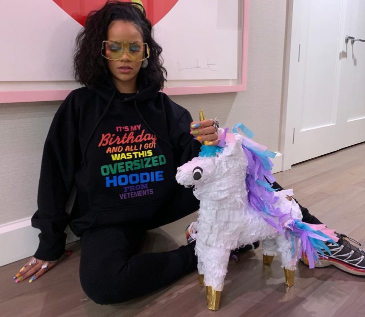 Cantante Rihanna vestida con hoodie grande, negro con letras del color del arcoíris y una piñata de unicornio, tendencia oversized