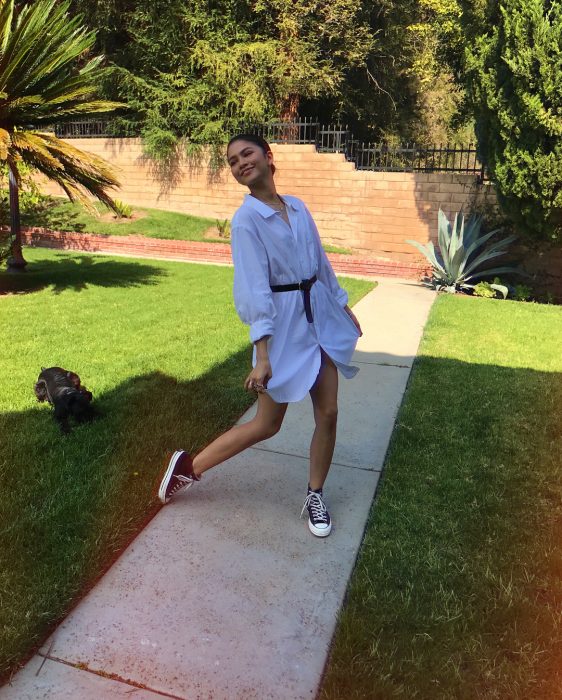 Zendaya modelando en jardín con camisa grande y cinturón negro como vestido, tenis converse, tendencia oversized