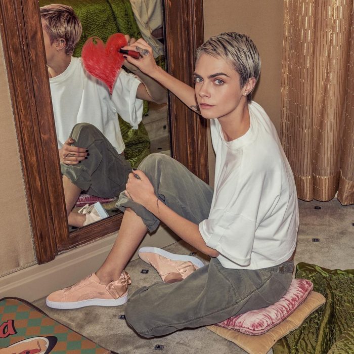 Actriz y modelo Cara Delevingne, chica pintando un corazón con labial en el eespejo, cabello rubio con corte pixie, playera blanca grande, pantalones grises anchos y tenis rosas durazno, tendencia oversized