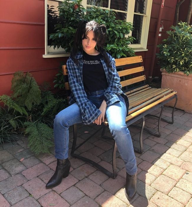 Singer Camila Cabello sitting on a wooden band, with high ponytails, blue jeans and large plaid shirt, oversized trend
