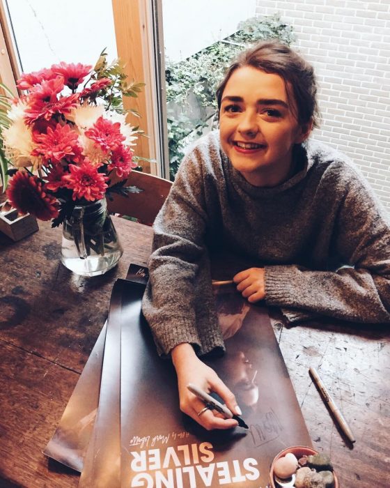 Actriz Masie Williams de Game of Thrones firmando autógrafos y sonriendo, vestida con un suéter grande tejido, tendencia oversized