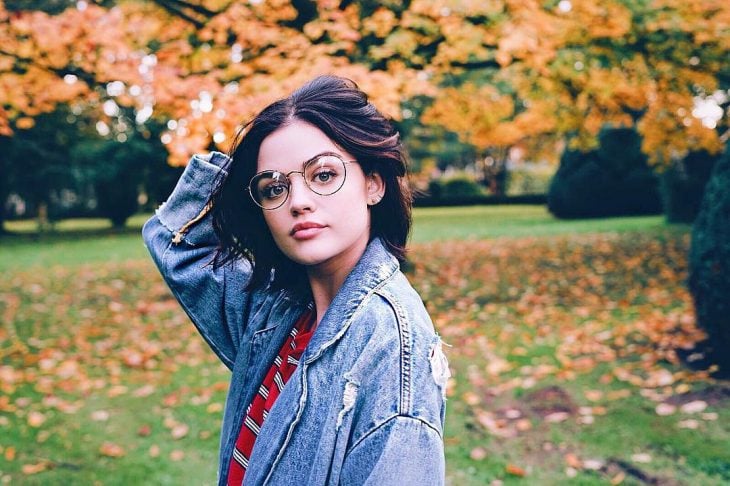 Actress Lucy Hale modeling outdoors in a park, wearing large magnifying lenses and a worn denim jacket, oversized trend
