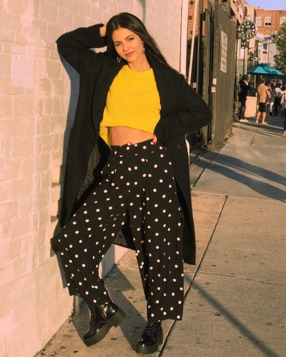 Actriz Victoria Justice, chica recargada en la pared usando un suéter amarillo mostaza, un saco negro, largo y grande y pantalones polka dots anchos, tendencia oversized