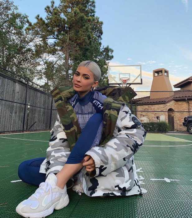 Kylie Jenner sitting on a basketball court, with gray hair, tennis and jacket with large military camouflage, oversized trend